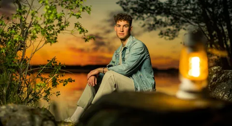 Nuori mies istuu järven rannalla auringonlaskun aikaan. 