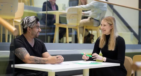 Kaksi opiskelijaa hymyilevät toisilleen istuessaan pöydän ääressä.
