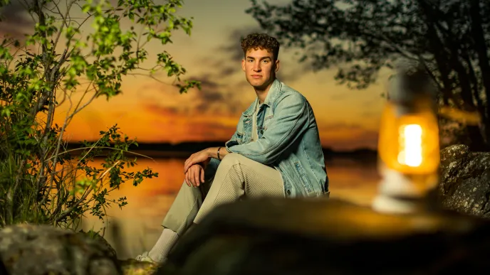 Nuori mies istuu järven rannalla auringonlaskun aikaan. 