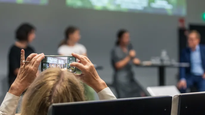 Yleisössä istuva nainen kuvaa puhujia auditoriossa älypuhelimella