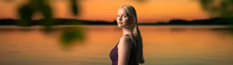 Henkilö seisoo järven rannalla auringon laskun aikaan.